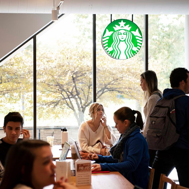 A group of Drake University students both standing and sitting at tables inside of the Starbucks location at Olmsted